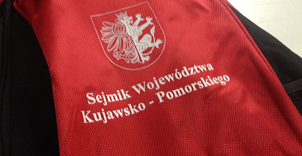 Biały nadruk Sejmiku Województwa Kujawsko-Pomorskiego na czerwonej odzieży